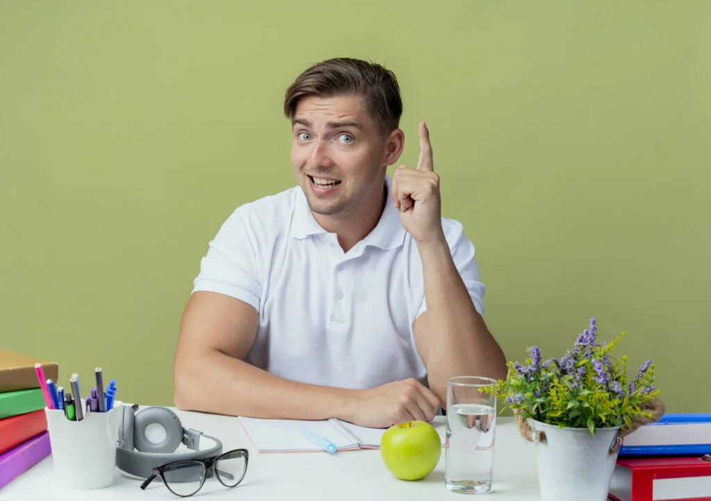 Student at study desk pointing up, surrounded by notebooks, pens, headphones, apple, water glass, and potted plant