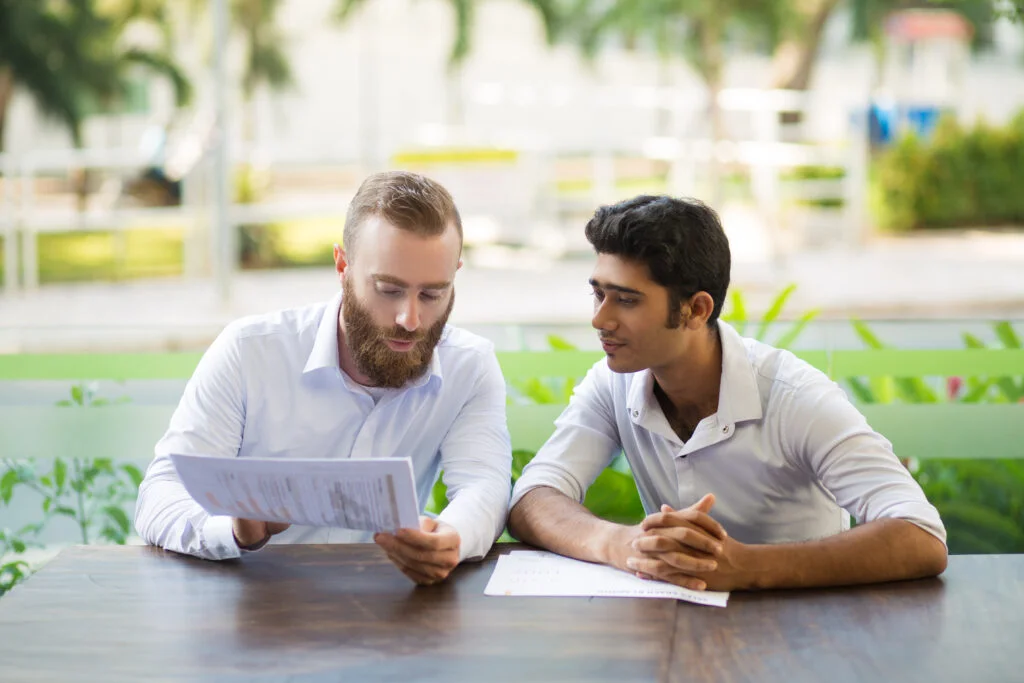 Two business professionals—one holding and reviewing a printed document, the other listening attentively—seated at a wooden table in an outdoor café setting.